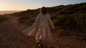 A woman in a coat walks on a scenic coastal dirt road as the sun sets, casting warm tones.