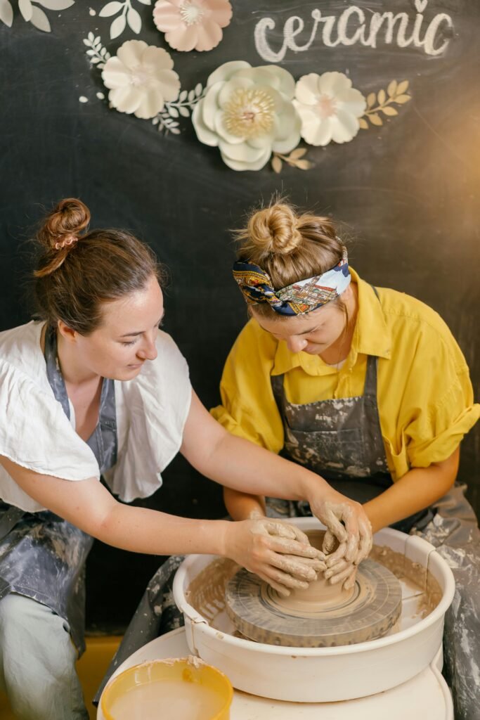 Two women engaging in a hands-on pottery workshop, shaping clay on a spinning wheel with handmade ceramic art in the background.